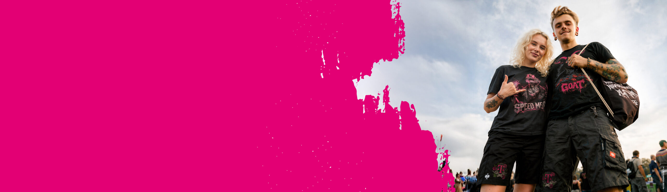 Zwei Personen, die schwarze Shorts und dazu passende schwarze T-Shirts mit Aufdruck tragen, stehen lächelnd nebeneinander bei einer Veranstaltung im Freien. Auf der linken Seite des Bildes ist ein großer Farbtupfer in leuchtendem Pink zu sehen.