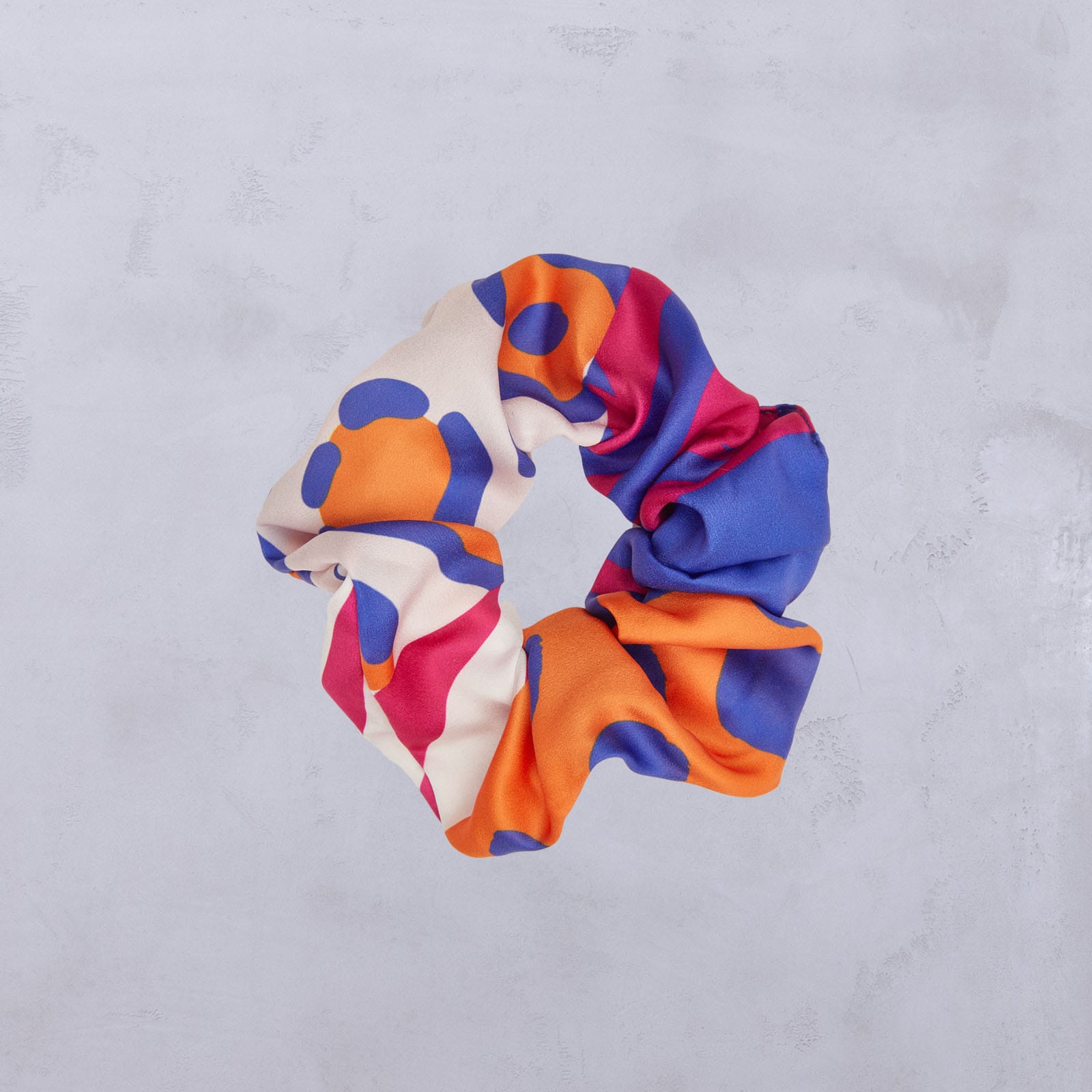 Ein farbenfrohes Stoff-Haargummi mit abstrakten Mustern in Orange, Blau, Creme und Magenta auf einem hellgrauen, strukturierten Hintergrund.