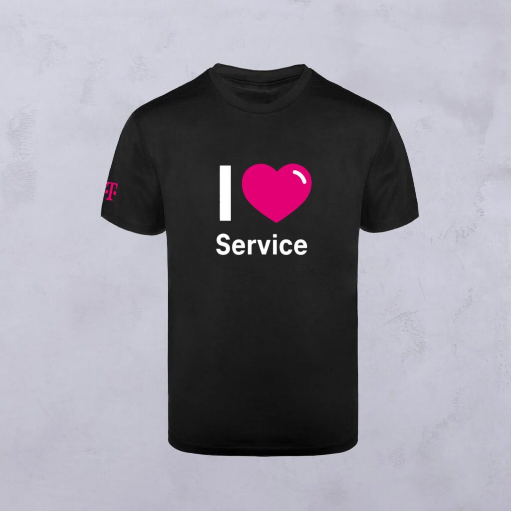 Schwarzes T-Shirt mit Ihrem Motiv – „I ♥ Service“ in weißer und rosa Schrift auf der Vorderseite sowie ein kleines rosa Logo auf dem linken Ärmel vor hellgrauem Hintergrund.