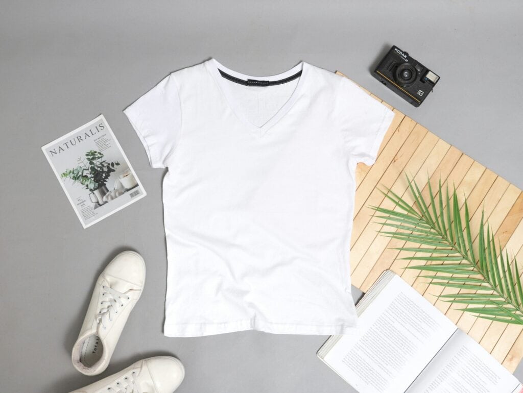 Ein schlichtes weißes T-Shirt mit V-Ausschnitt liegt auf einer grauen Fläche, um das herum weiße Turnschuhe, eine Kamera, eine Pflanzenzeitschrift, ein aufgeschlagenes Buch, ein Palmblatt und eine Bambusmatte angeordnet sind.