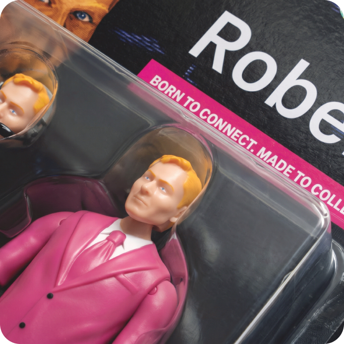 Nahaufnahme einer verpackten Actionfigur in rosa Anzug und Krawatte, mit heller Haut und blonden Haaren. Auf der Verpackung steht der Text „Born to connect. Made to collect.“ und ein Teil des Namens „Robe“ ist zu sehen, was auf den vollständigen Namen der Figur, RoberT, hindeutet.