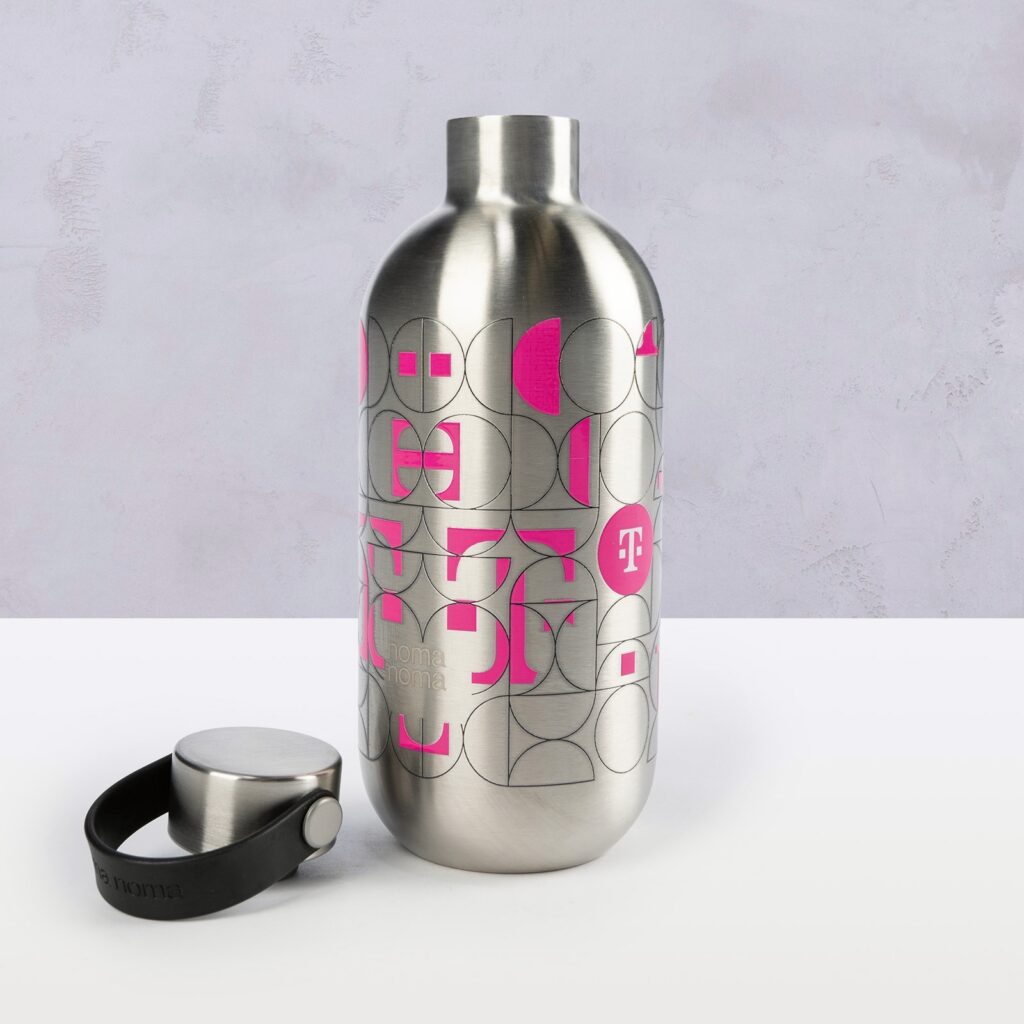 Eine RIDDLE T-Flasche mit einem Korpus aus Edelstahl, einem abnehmbaren Deckel und verziert mit rosa geometrischen Formen und Mustern steht auf einer weißen Fläche vor einem hellgrauen Hintergrund.