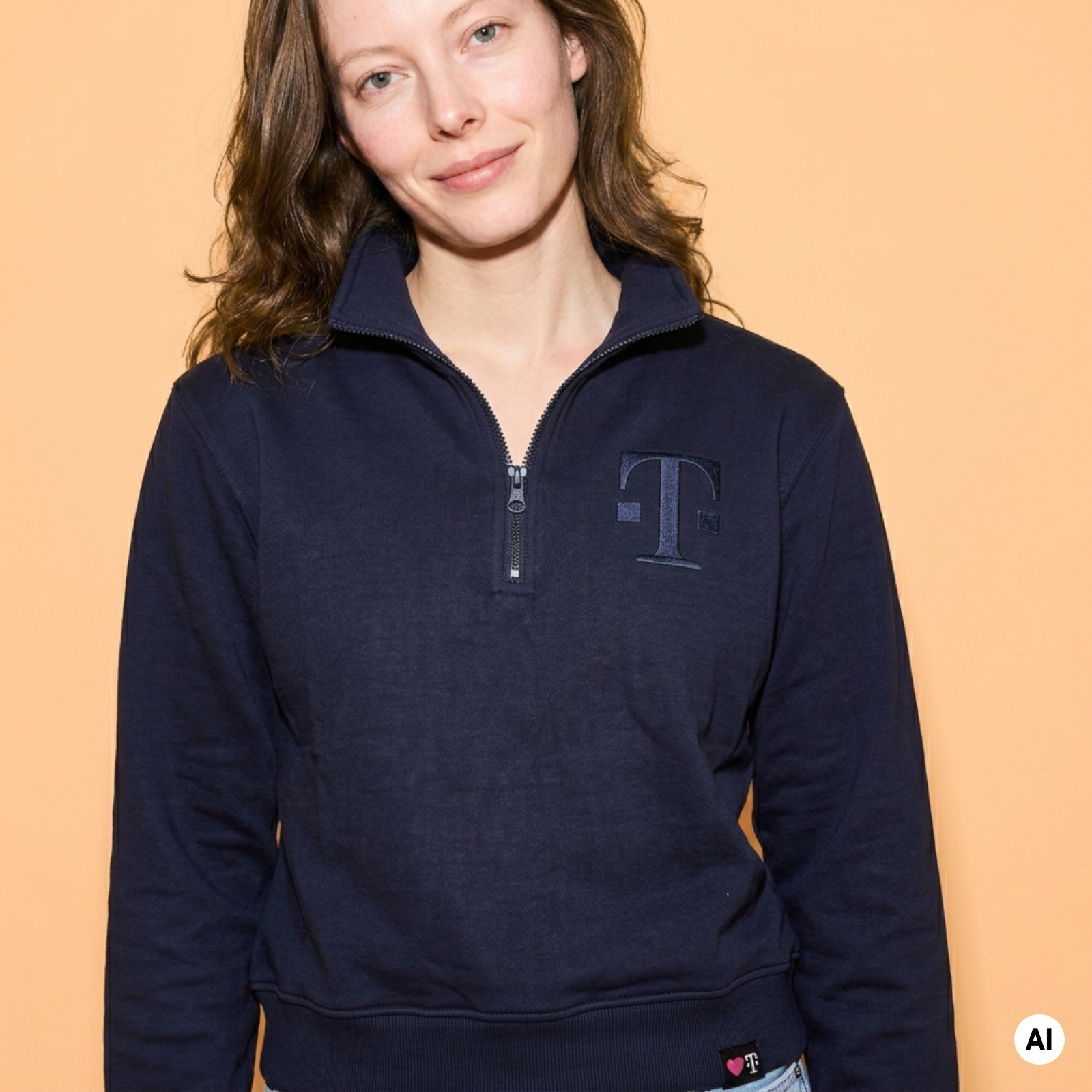 Eine Frau mit heller Haut und braunen Haaren, die ein marineblaues Sweatshirt mit Reißverschluss und einem T-Logo trägt und vor einem pfirsichfarbenen Hintergrund steht.