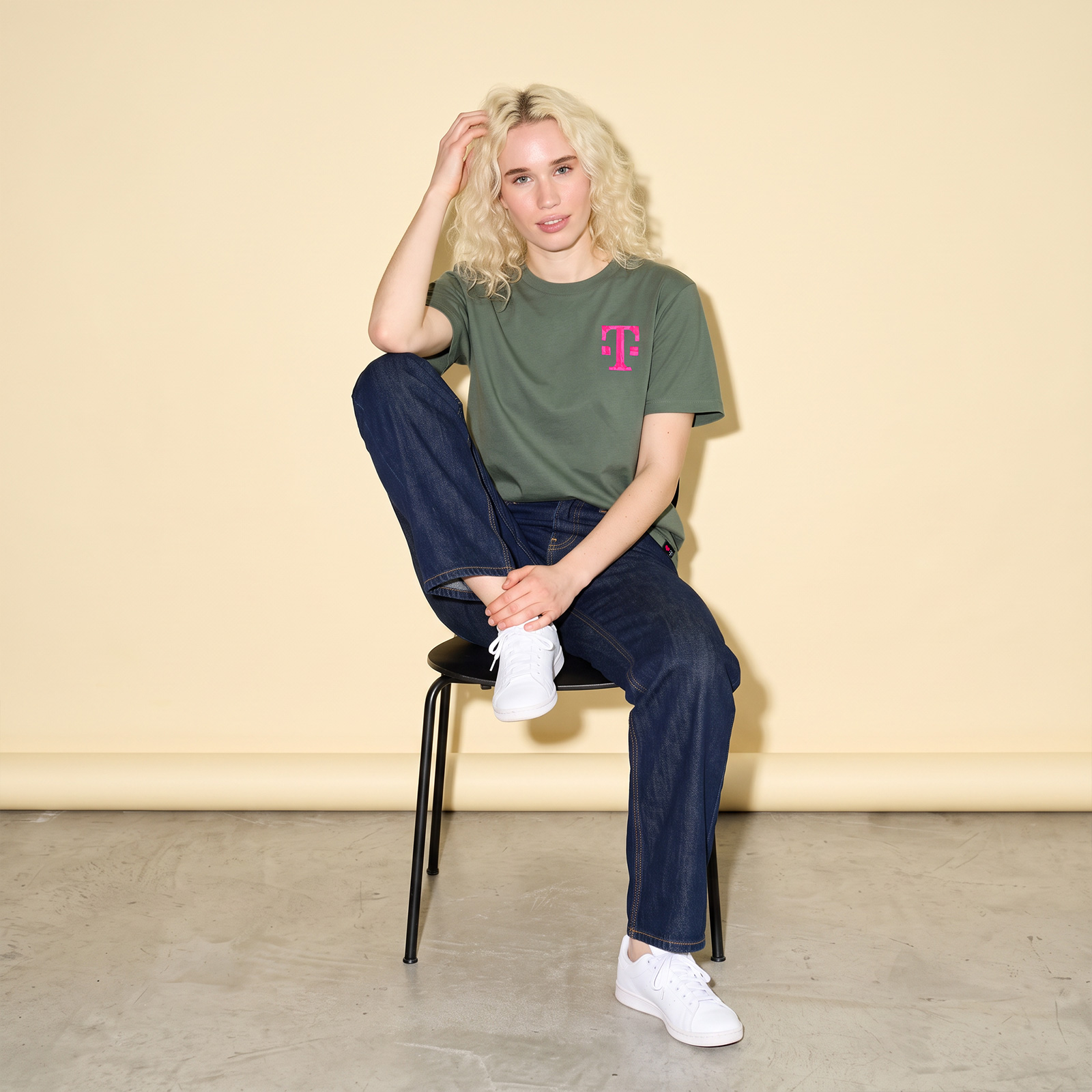 Eine Person mit blonden Haaren sitzt auf einem schwarzen Stuhl und trägt ein grünes TAPE ART-T-Shirt mit einem rosa T-Mobile-Logo, eine dunkle Jeans und weiße Turnschuhe.