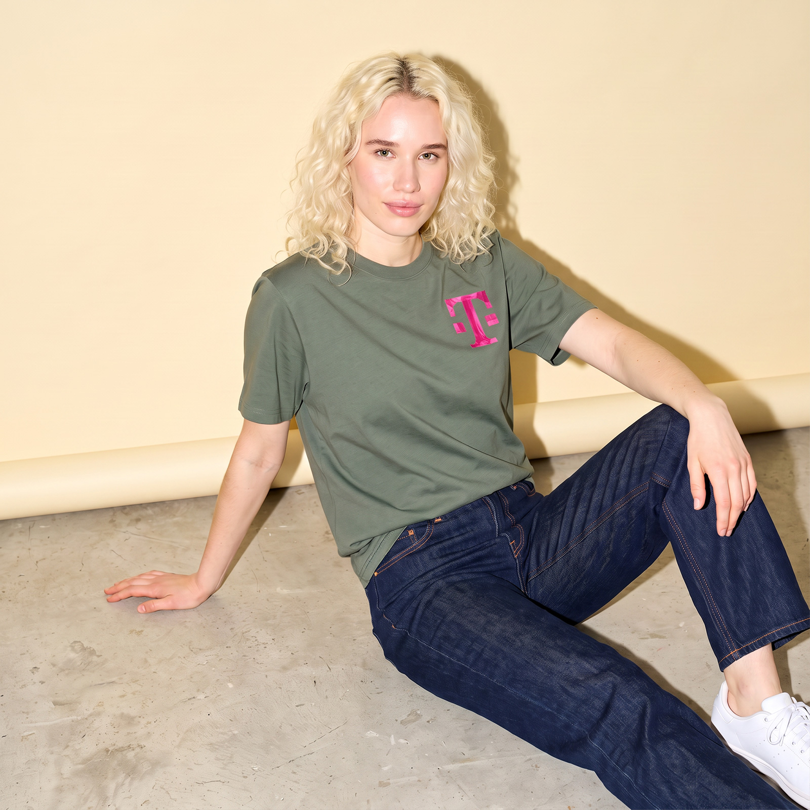 Eine Person trägt ein olivgrünes TAPE ART-T-Shirt mit einem rosa T-Logo auf der Brust, dazu dunkelblaue Jeans und weiße Sneaker.