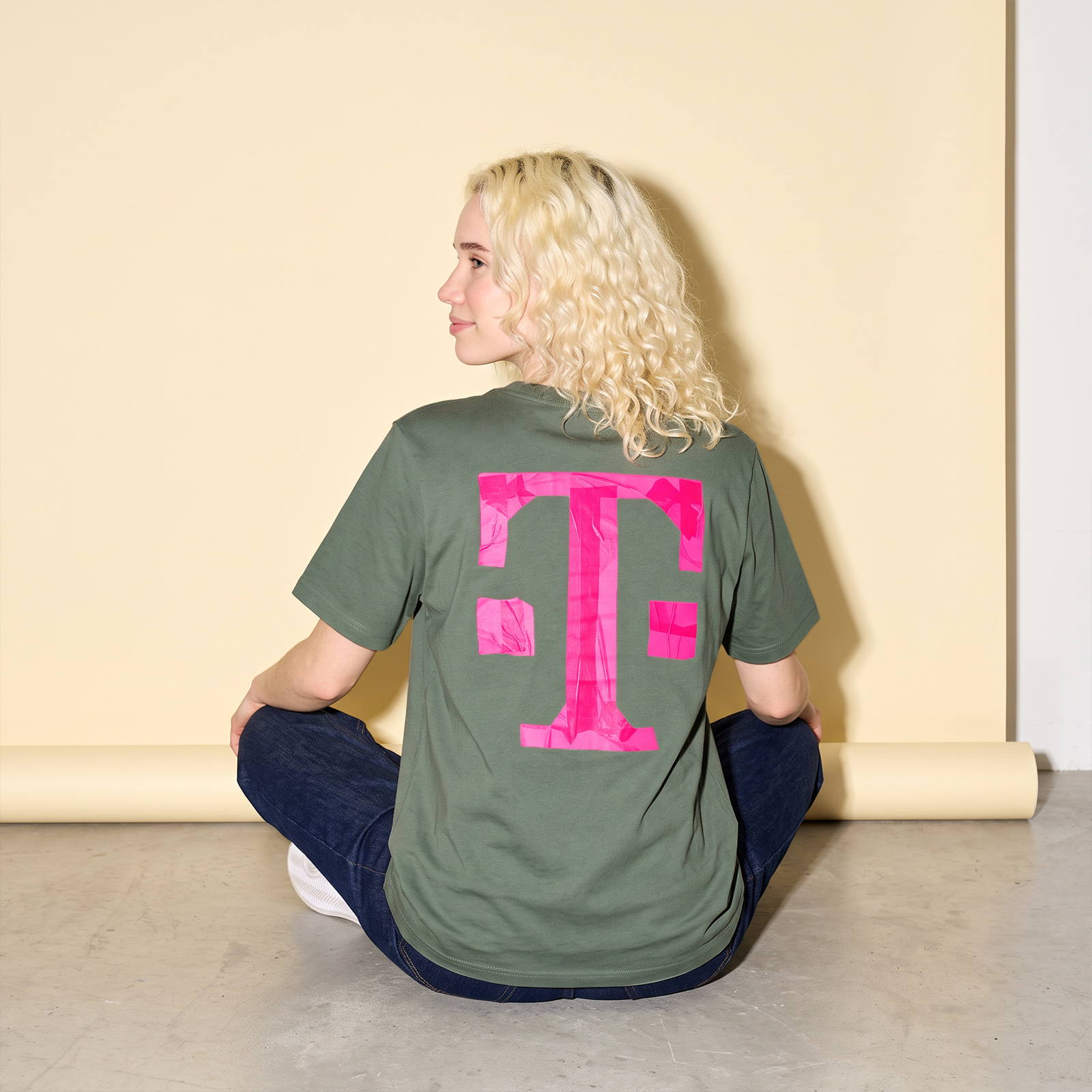 Eine Person sitzt im Schneidersitz und trägt ein olivgrünes TAPE ART-T-Shirt, auf dessen Rücken ein großes rosa T aufgedruckt ist.