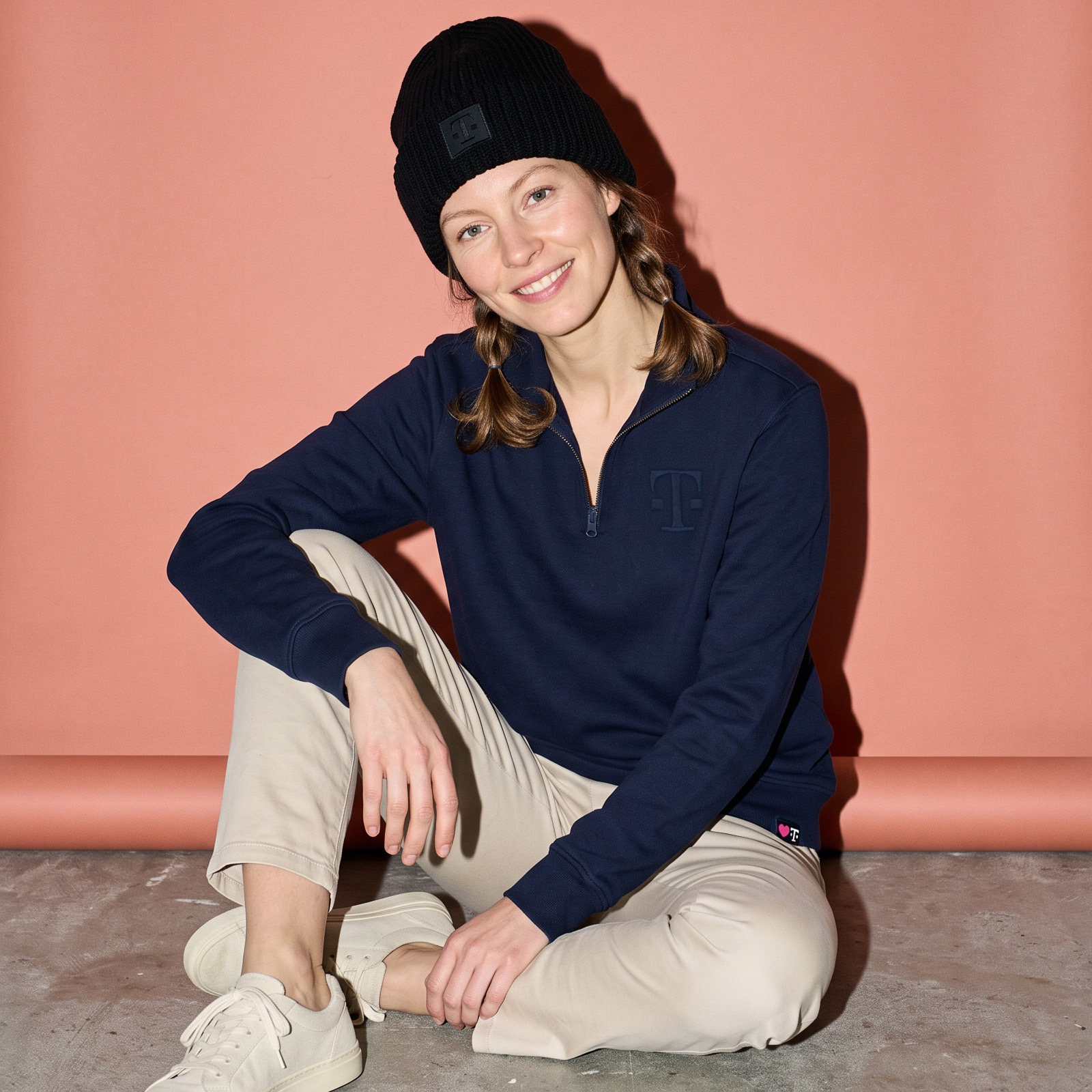 Sitzende Person in dunkelblauem Half-Zip-Sweater trägt die LOGO BEANIE BLACK vor roséfarbenem Hintergrund