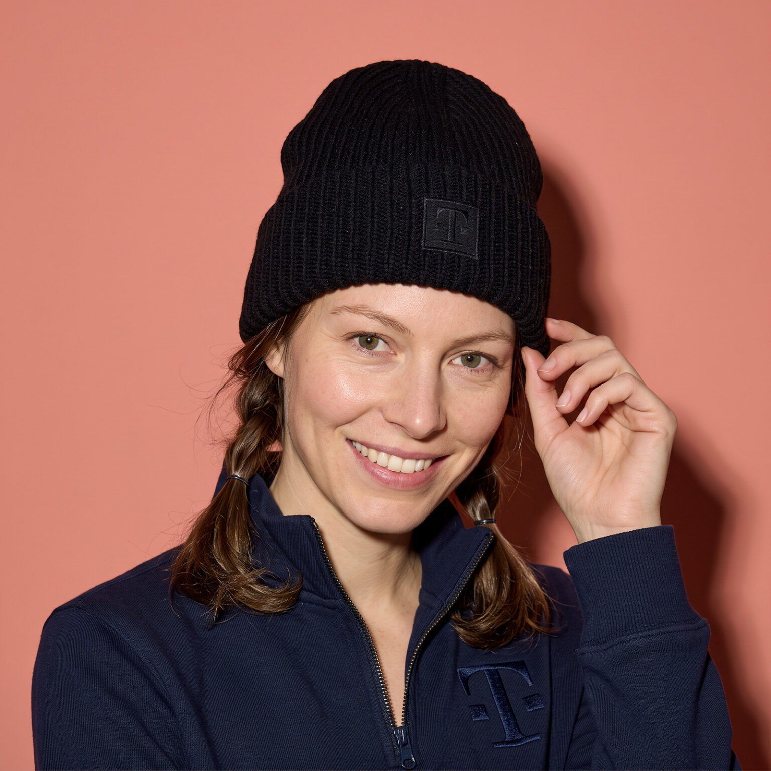 Lächelnde Person trägt die LOGO BEANIE BLACK vor roséfarbenem Hintergrund